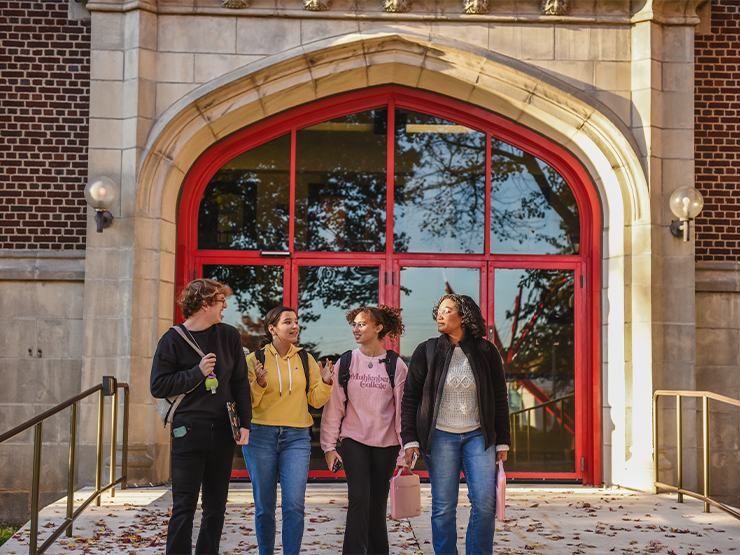 Four students speak with one another as they walk away from a large arched doorway, leaves scattered on the sidewalk before them.